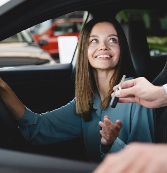 A happy female customer is sitting inside the car at the wheel, while a male assistant is giving her the car keys
