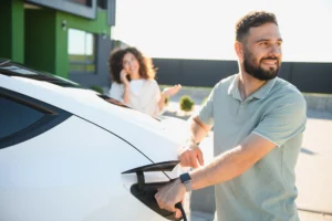 A man charging a white electric vehicle at home while his partner talks on the phone, illustrating how to save on fuel costs with EV finance options.