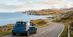 A blue electric Fiat 500 drives along a winding coastal road with views of the ocean and rocky cliffs.