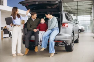 Family buying new car at SUV dealership showroom, saleswoman explaining features with laptop, happy customers choosing vehicle