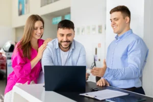 Young couple reviewing information on laptop with car sales consultant at dealership desk, comparing vehicle options, types such as electric, petrol, hybrid, prices, financing plans, and purchase details together